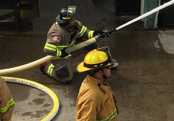 one firefighter crouches on the ground using a hose and another watches