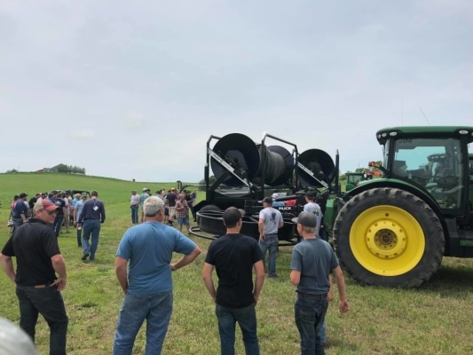 a crowd in a field gathered around a tractor and turn table reel hose cart
