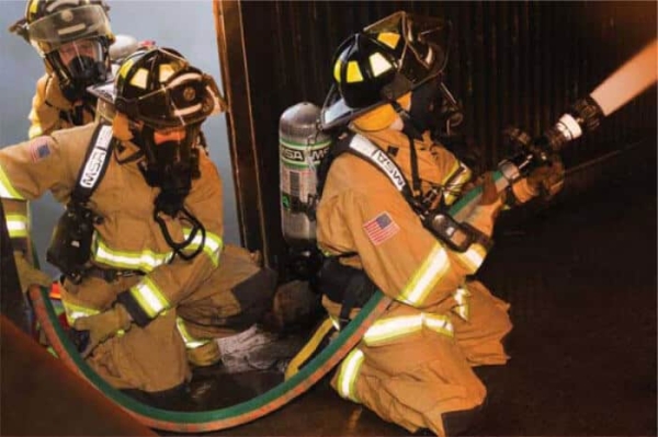 three firefighters crouched and holding a Hi-Combat II hose in the entryway of a building as they put out a fire in training