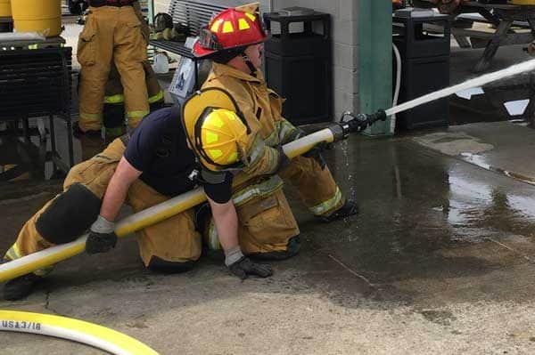 The backup firefighter kneels behind the nozzle operator of the ground for support, displaying the proper form for acting as support in hose handling.