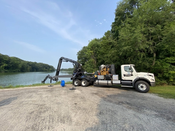 a boom truck parked and stabilized next to water with a blue hose rolled up next to the truck and ready to use