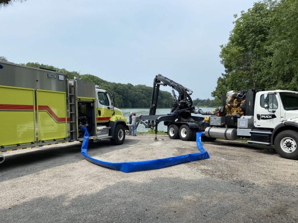 a fire truck and boom truck connected via a blue hose while parked next to the water