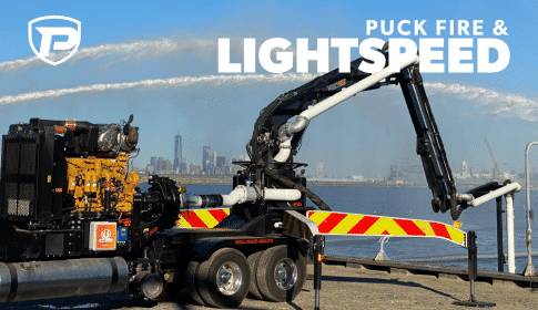 Puck Fire & LightSpeed; a Puck FOrce Feed truck with Lightspeed control panel has extended boom in water and hoses from a nearby Quick Attack shooting water overhead in the background
