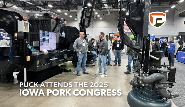 the Puck booth at Iowa Pork Congress in the Iowa Events Center with a large black force feed trailer with extended hydraulic boom is parked and people around it talk in conversations