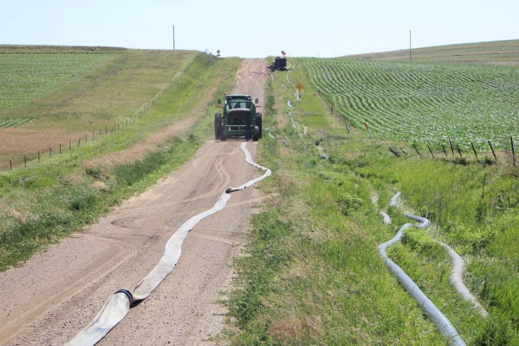 a green Puck HC 8 laying hose while the towing tractor pulls it down a dirt road. a booster pump and inflated hose lay along the ditch next to the fields
